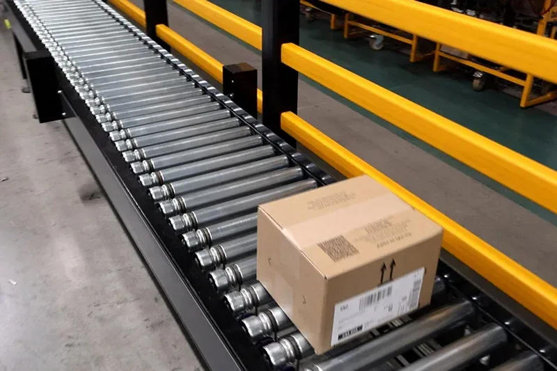 Transportador de rodillos de eje recto que transporta una caja de cartón junto a barreras protectoras.